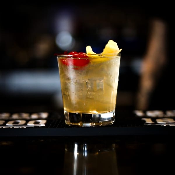 A cocktail with ice, cherries, and lemon peel sits on a dark bar surface.