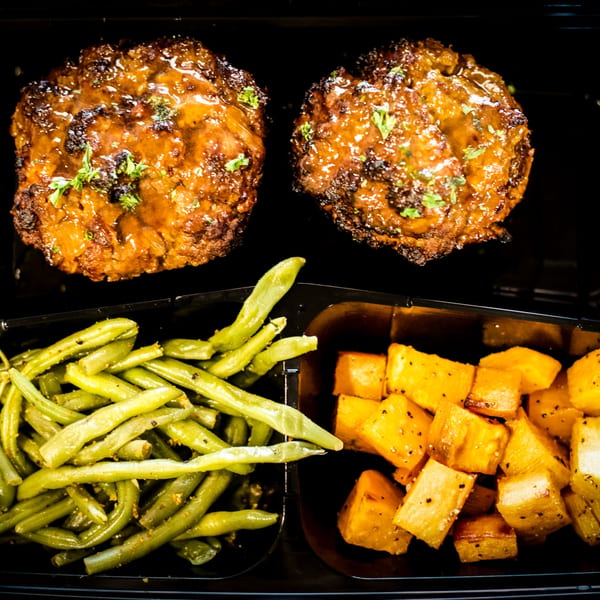 Two glazed meatloaves with green beans and roasted potatoes in a black meal prep container.
