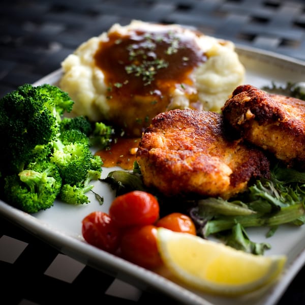 A plate of golden-brown crab cakes, mashed potatoes with gravy, broccoli, and cherry tomatoes.