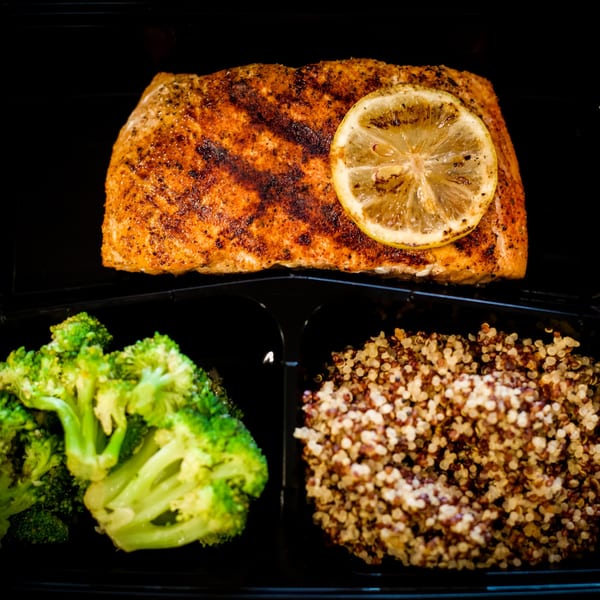 Seasoned salmon with a lemon slice, broccoli, and quinoa in a black meal prep container.