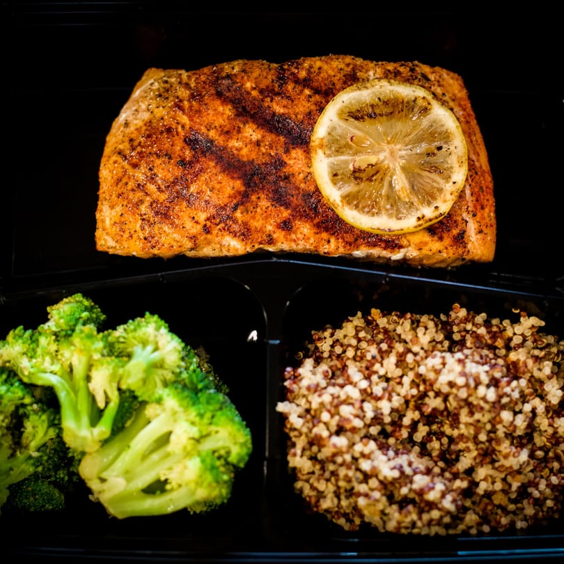 Seasoned salmon with a lemon slice, broccoli, and quinoa in a black meal prep container.