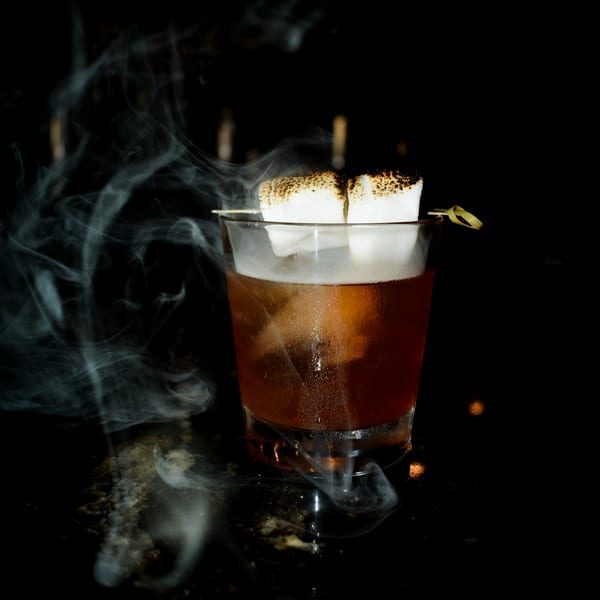 Smoky cocktail with toasted marshmallows on a skewer, against a dark background.