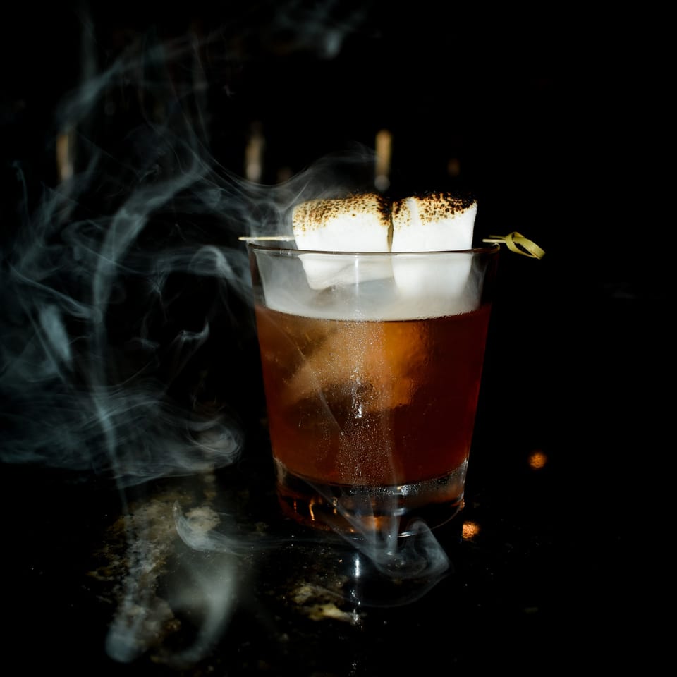 Smoky cocktail with toasted marshmallows on a skewer, against a dark background.