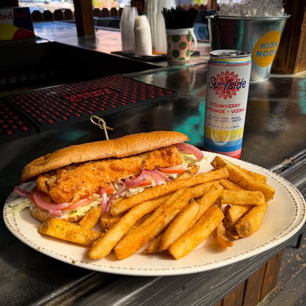 A fried fish sandwich with lettuce, tomato, and red onion, served with fries and a Surfside Strawberry Lemonade + Vodka can.