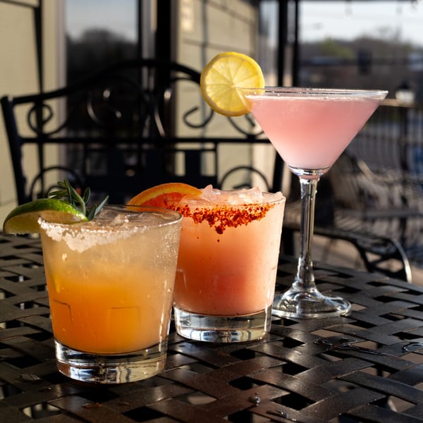 Three colorful cocktails, garnished with citrus and herbs, sit on a dark patio table.