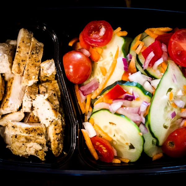 Sliced chicken breast and a colorful salad with cucumbers, tomatoes, and onions in a black container.