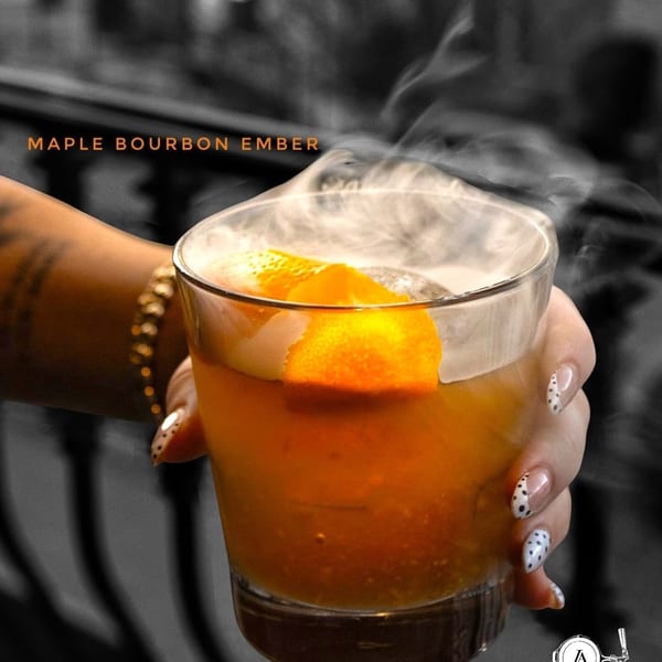 Hand holding a smoking Maple Bourbon Ember cocktail garnished with an orange peel.